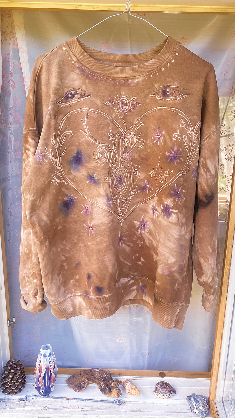 A brown tie-dye long-sleeve sweatshirt with a heart and eye graphic design, hanging in a display case with decorative items on a shelf below.